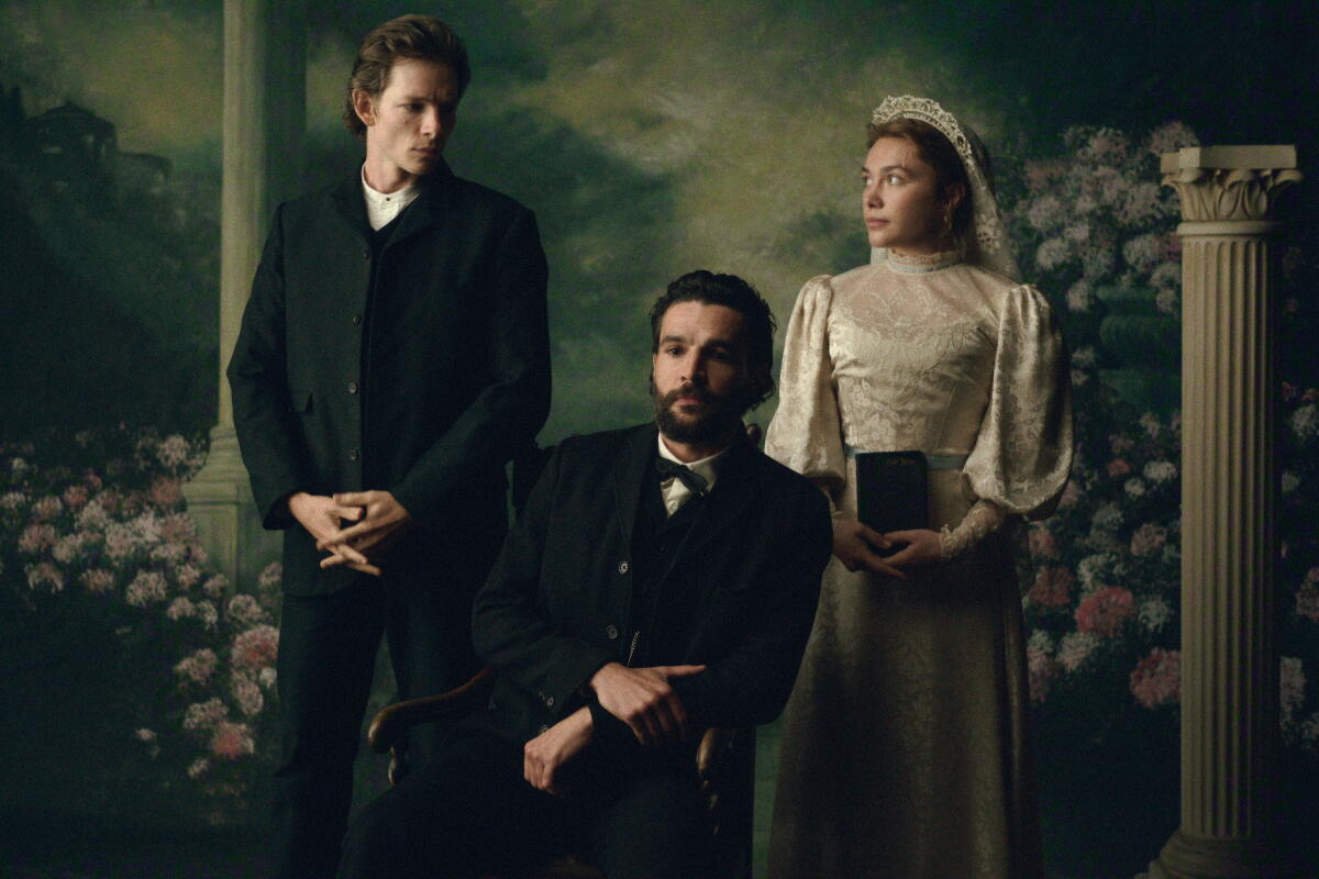 A man in a dark suit and a woman in a white bridal gown stand behind a man in a dark suit sitting in a chair.