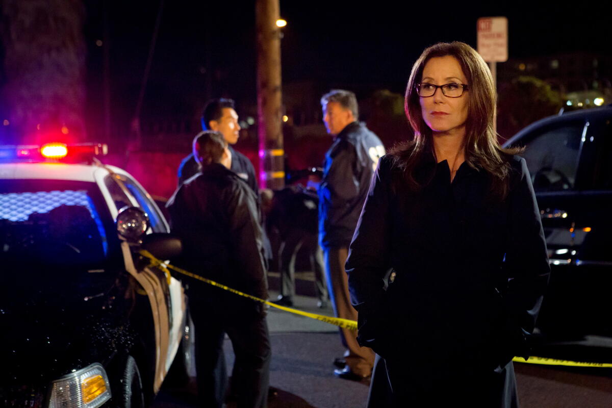 A bespectacled woman in a coat stands beside a cop car with its emergency siren lit