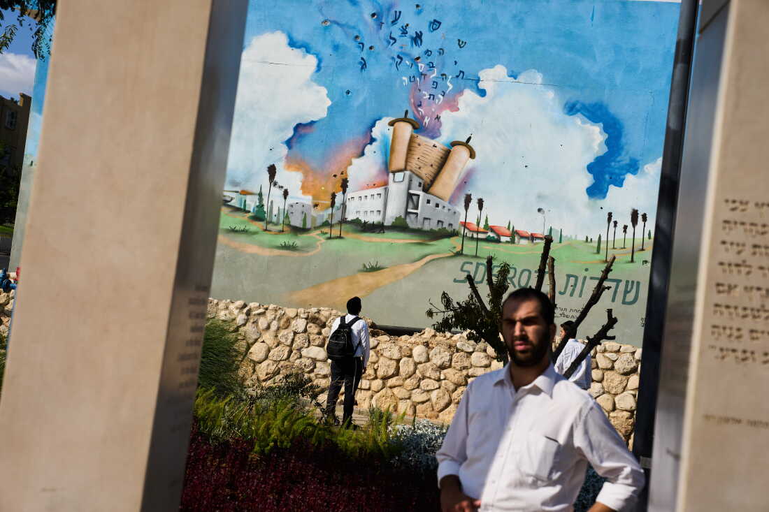 Three men visit a memorial at a police station attacked by Hamas on October 7, 2023, in Sderot, southern Israel, on October 4, 2026. The memorial includes a large mural of a white building with a giant Torah scroll on top of it.
