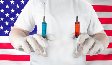 Medical worker in gloves holding syringes with blue and red liquid against American flag backdrop.