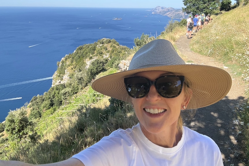 Jennifer Tucker smiling, wearing a wide brimmed hat, sunglasses, white t-shirt, doing a selfie on a hike.