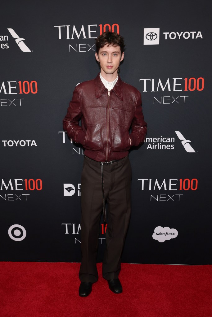 Troye Sivan attends the Time100 Next Gala in a burgundy leather jacket, white shirt, and dark trousers on a red carpet.