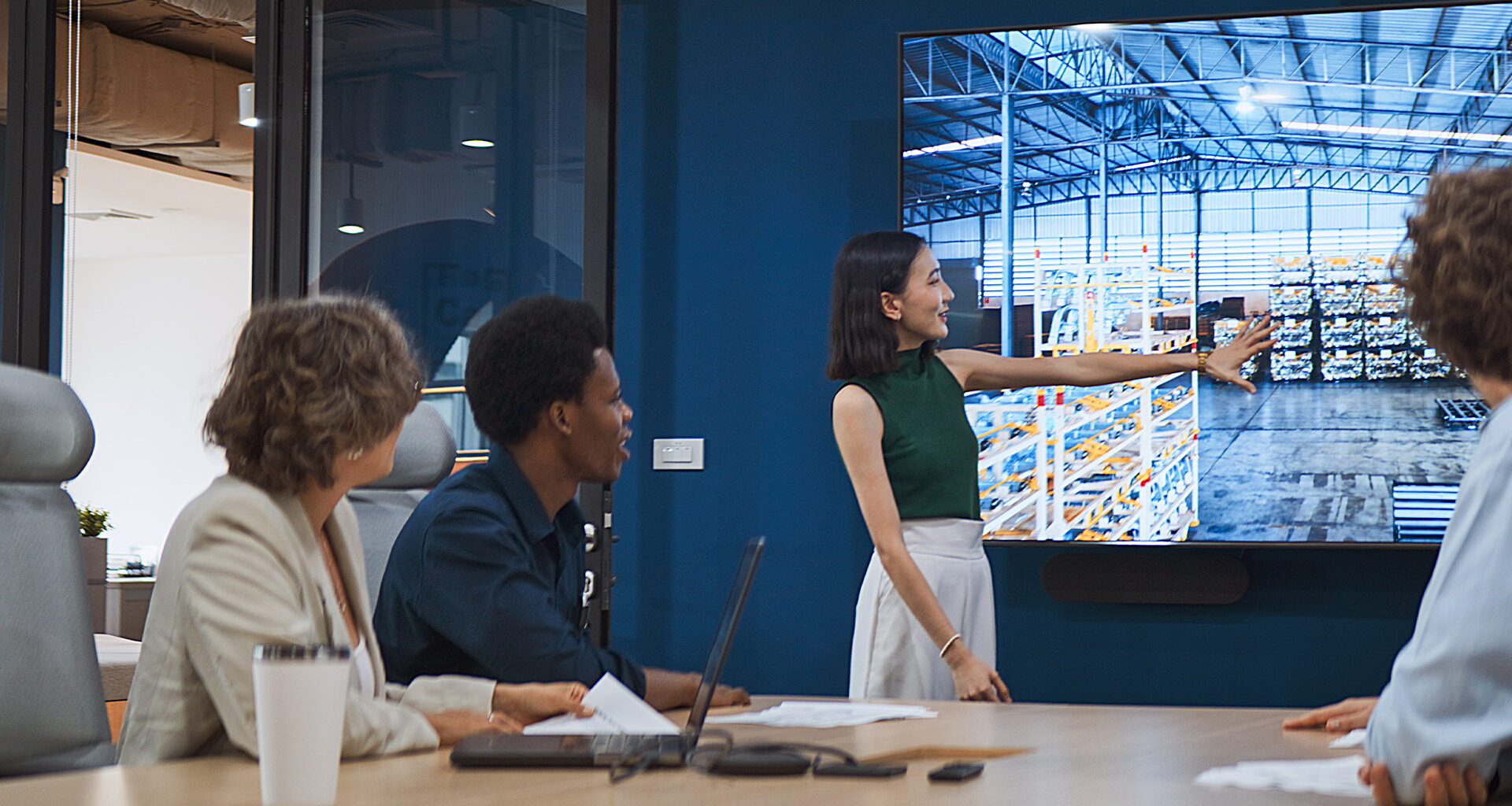 Four people in a conference room watch a presenter point to a warehouse scene on a large display.