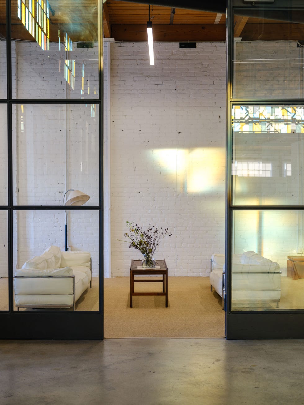 Modern interior with glass walls, white couches, a wooden table, and decorative flowers under warm lighting.