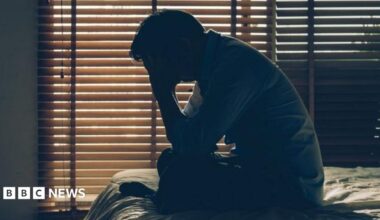 A man sits crossed legged on a bed in front of closed window blinds with his head in his hands. He is in silhouette.