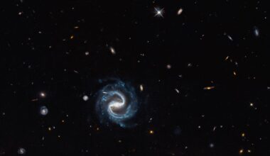 A spiral galaxy with two distinct arms forming a backwards "S" shape is in the foreground, against a background of many other much smaller, more distant galaxies. In the right lower corner, a gravitational lens can be seen, with a large, distinctive arc to the upper left.
