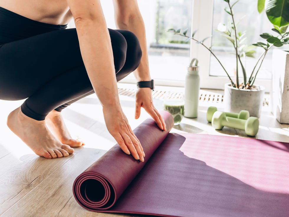 Close up of young woman practicing yoga, exercising at home. Unrolling yoga mat, getting prepare to work person rolling up a purple yoga mat next to plants and dumbbells.