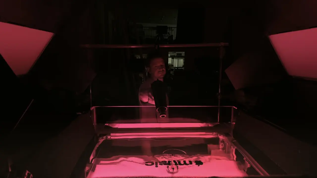 Person sitting at a desk illuminated by bright pink neon light, with rippling patterns visible in mineral oil below, demonstrating the sound-to-visual process used to create Apple Music's new logo