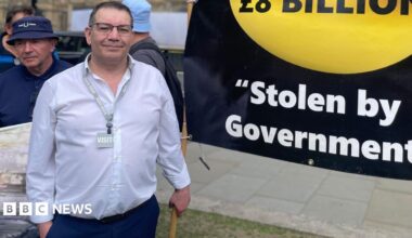 Picture of a middle aged white man, in a white shirt standing next to a black and gold banner stating: "Saved by Miners. I.O.U £8 billion. Stolen by Government".
