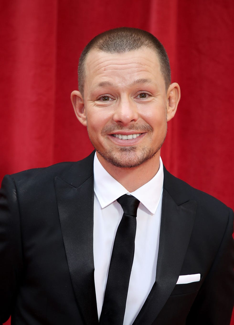 man in a suit smiling against a red curtain background.