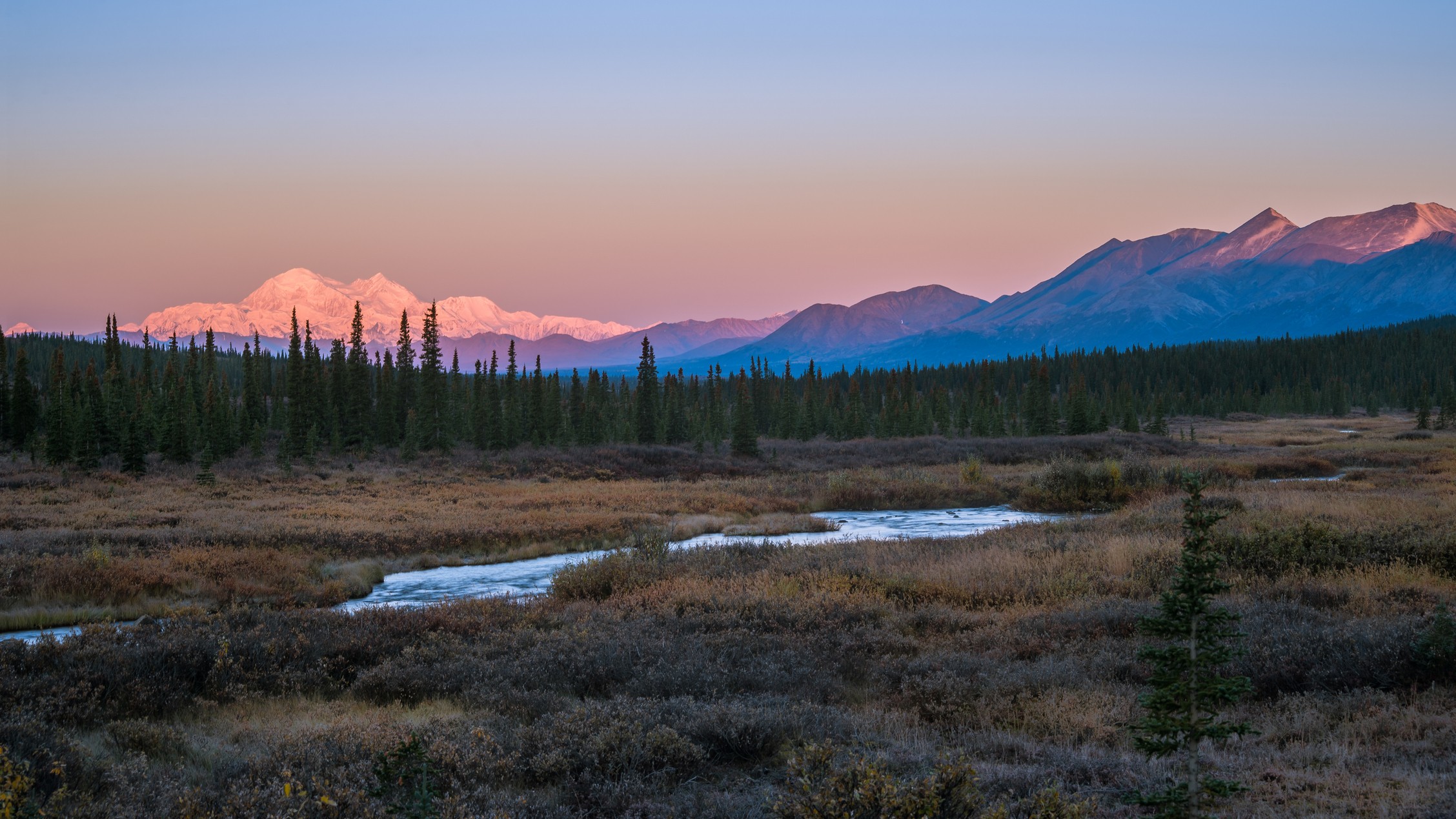 a lake in the foreground with trees in the background and mountains in the background, with a hint of pink.