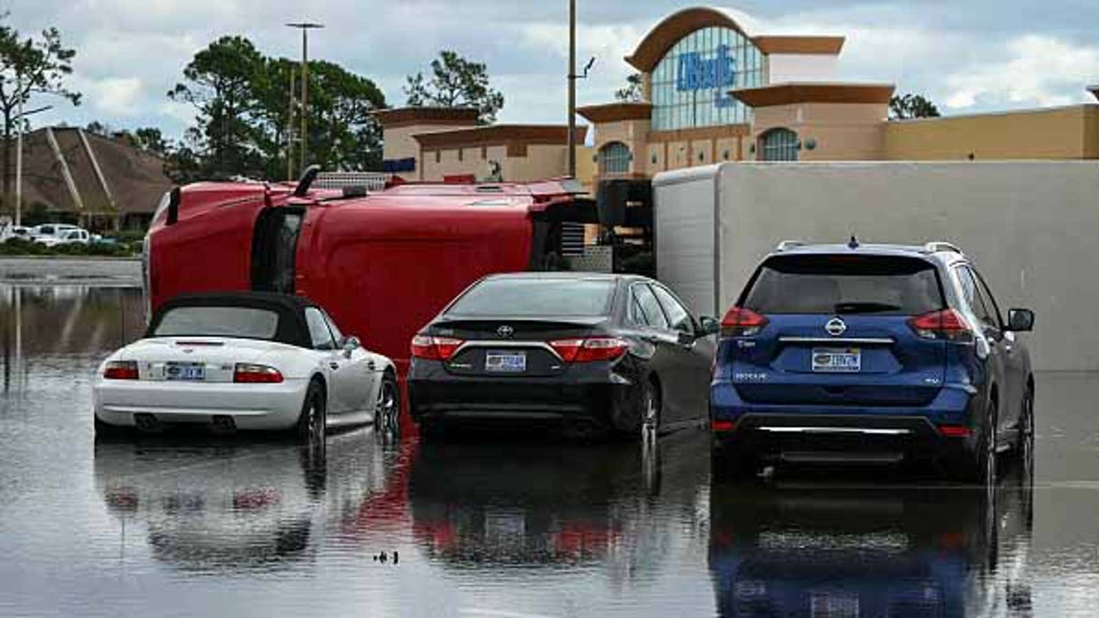 Flooded parking lot with an overturned red truck beside parked cars in front of a Publix store