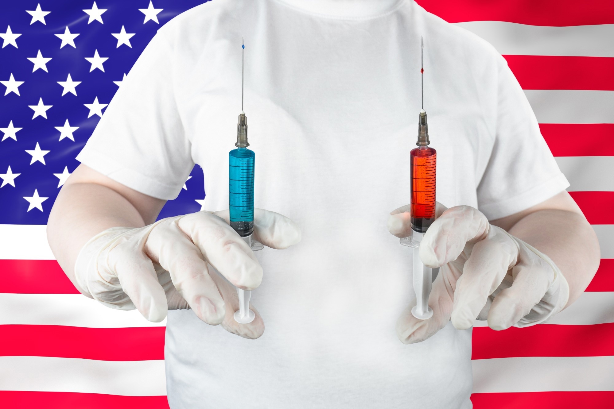 Medical worker in gloves holding syringes with blue and red liquid against American flag backdrop.