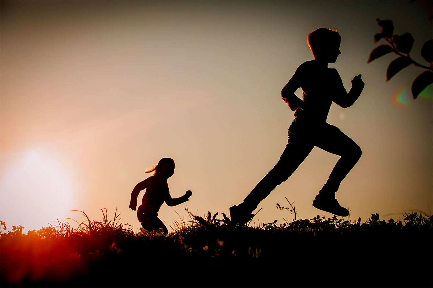 Silhouettes of two children running across a grassy hilltop at sunset.