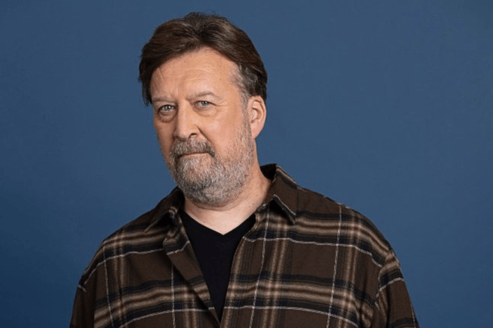 Ross Wilkes Ross Wilkes, stood against a dark blue backdrop for Coronation Street