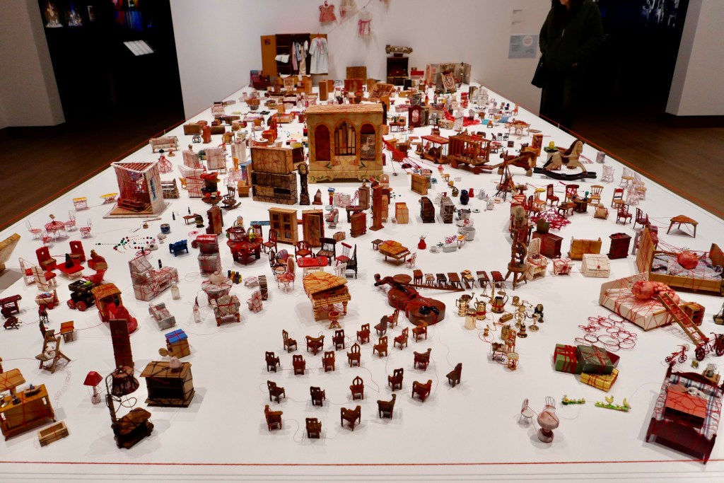 A large white table covered in miniature dollhouse furniture and other old flea market finds, all covered in red thread.