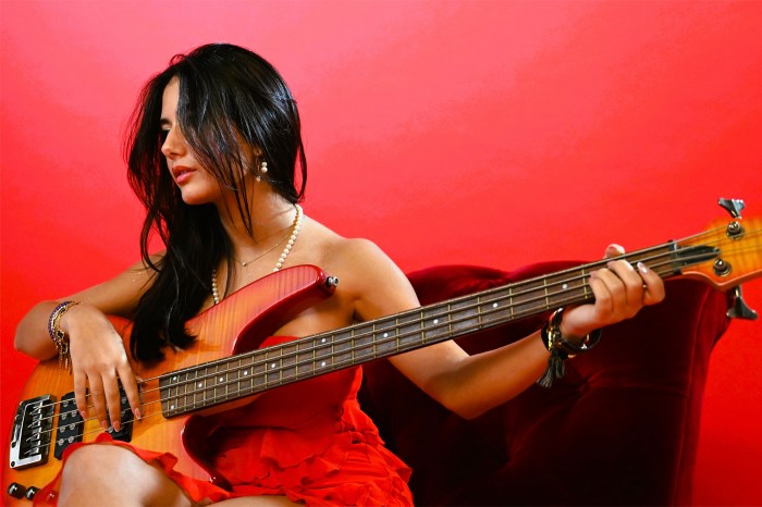 Feriani, in a red dress and pearl necklace, plays an electric bass while seated on a red velvet chair against a red backdrop.