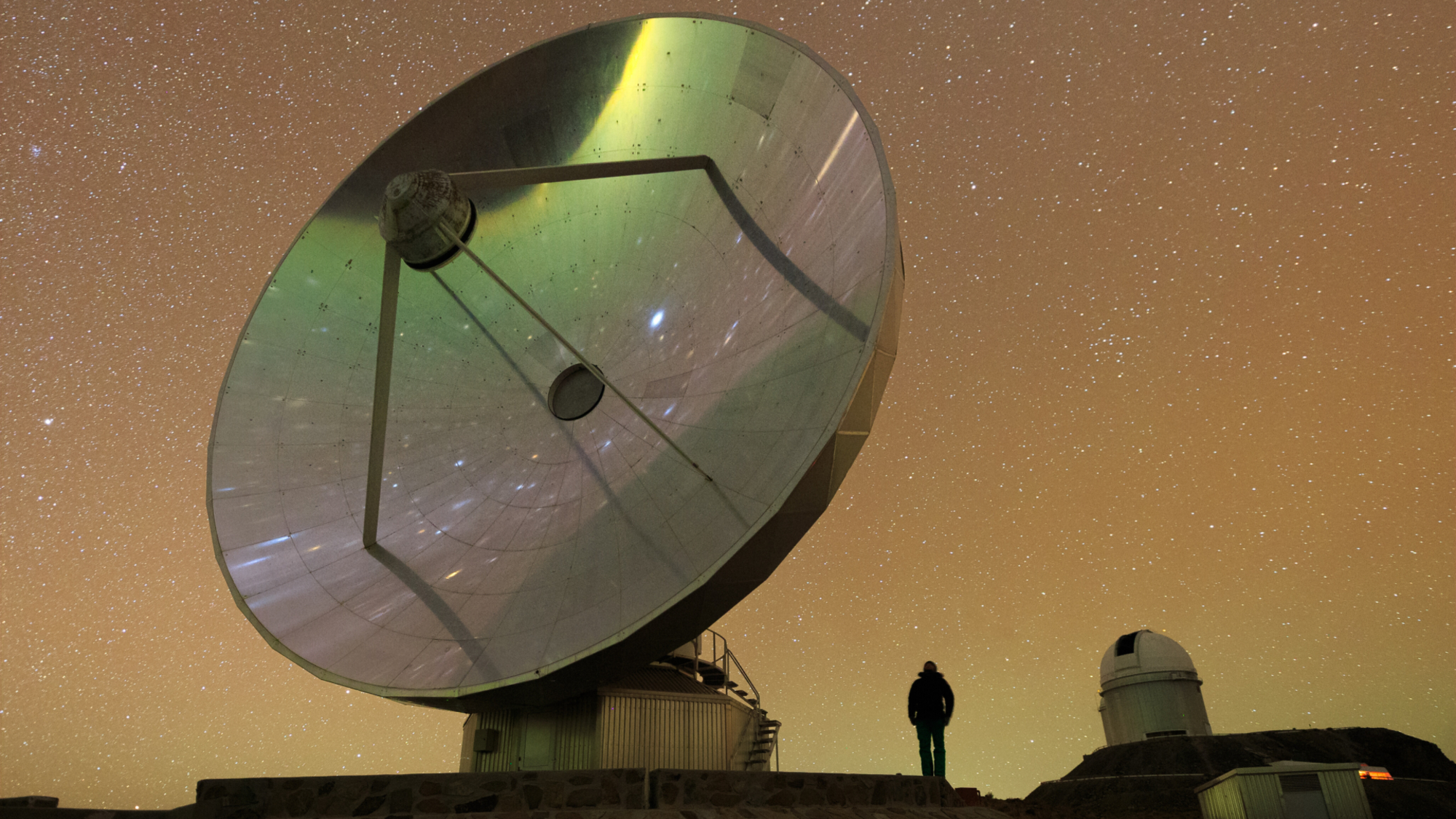A large telescope dish points skywards against the backdrop of a starry night sky infused with an orange glow. A man stands in the foreground and stars can be seen reflected in the telescope dish.