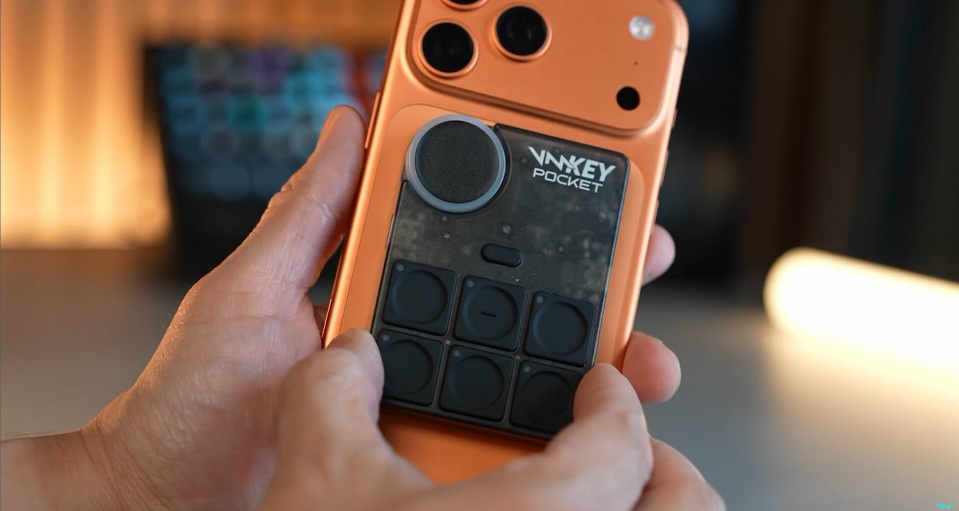 A hand holds an orange phone and places a black gadget on the back. The black gadget has six physical buttons and a volume wheel on the top left.