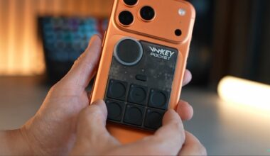 A hand holds an orange phone and places a black gadget on the back. The black gadget has six physical buttons and a volume wheel on the top left.