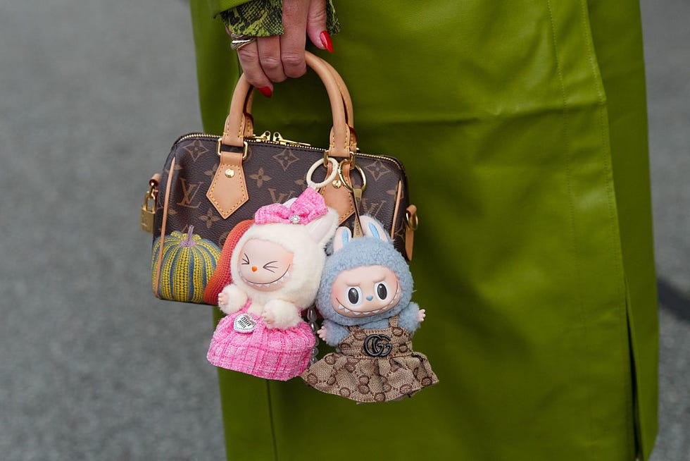 copenhagen, denmark august 07: a guest wears a bright olive green leather coat with a green snake print cuff peeking from the sleeve, a silver toned ring, and a glossy red pointed manicure, while a brown louis vuitton monogram coated canvas mini top handle bag with tan vachetta leather rolled handles, leather tabs, a gold tone zipper and padlock, and a yellow and orange pumpkin motif is carried; two labubu plush bag charms are attached one cream plush figure in a pink tweed dress with a matching bow, the other blue gray plush with bunny ears in a brown gucci gg monogram dress finished with a black double g emblem and gold tone hardware attachments, outside rotate, during copenhagen fashion week, on august 07, 2025 in copenhagen, denmark (photo by edward berthelot/getty images)