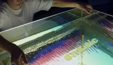 A person gestures over a shallow tray containing mineral oil with colourful sound wave patterns and a musical note created by vibrations from a transducer