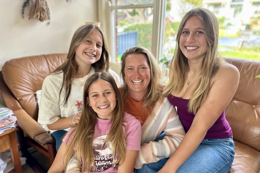 Three smiling teenaged girls sit cosily around their mum in a loungeroom.