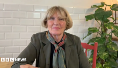 Denise Mackay with blonde hair, wearing an olive blazer and a pashmina. She is sitting at a wooden table on a red chair looking at the camera.