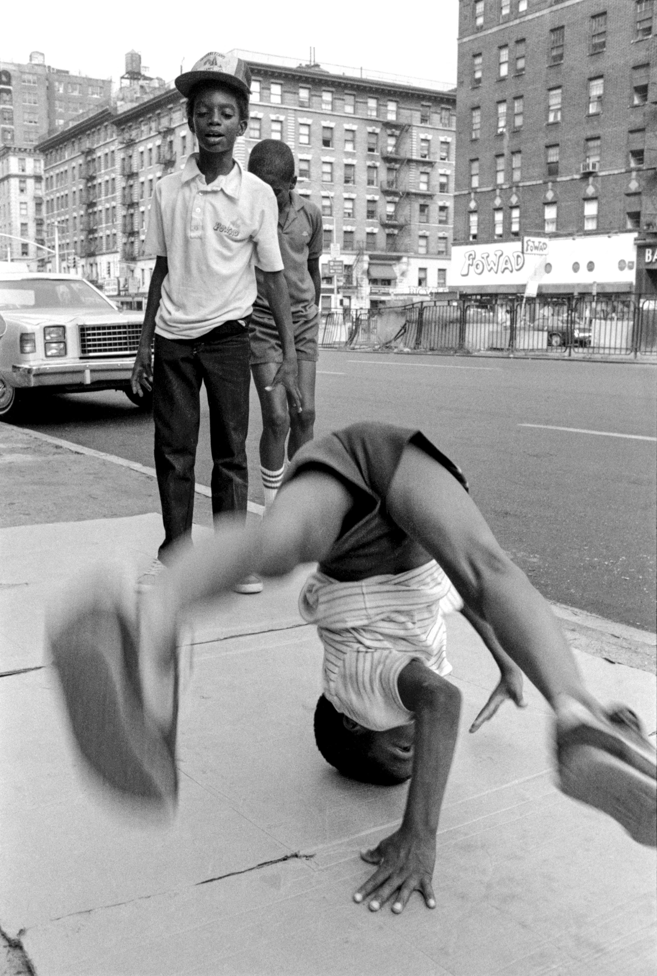 B boy breakdancing on a New York street with onlookers and tall buildings in the background, photographed by Henry Chalfant
