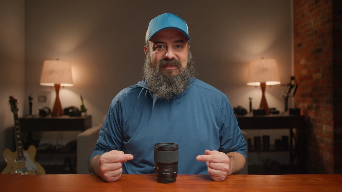Bearded man in a blue cap and blue shirt pointing at a black camera lens standing on a wooden table