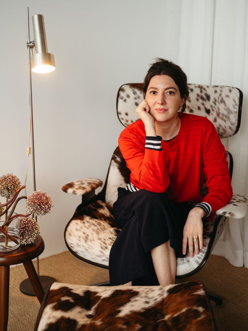 Person sitting in a stylish chair, wearing a red sweater, with a focused expression in a cozy indoor setting.