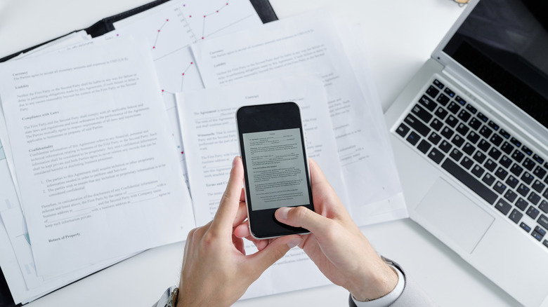 a person uses a smartphone to scan documents from a binder, macbook visible on desktop nearby
