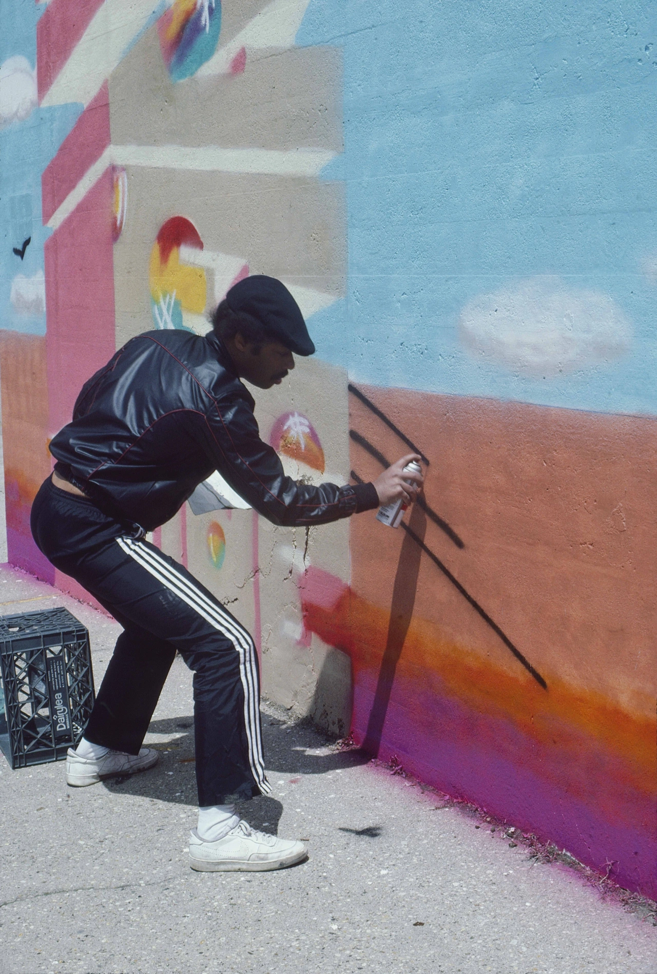 Artist in black tracksuit and cap spray-painting colourful abstract mural on urban wall against blue sky