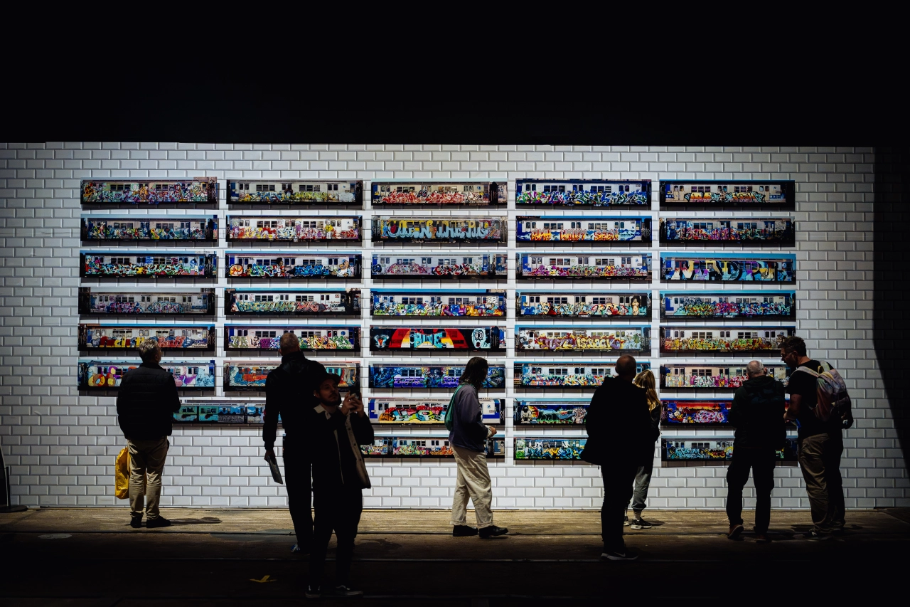 Silhouetted figures stand viewing a large wall installation of multiple horizontal panels displaying colourful street art and graffiti photographs on white brick