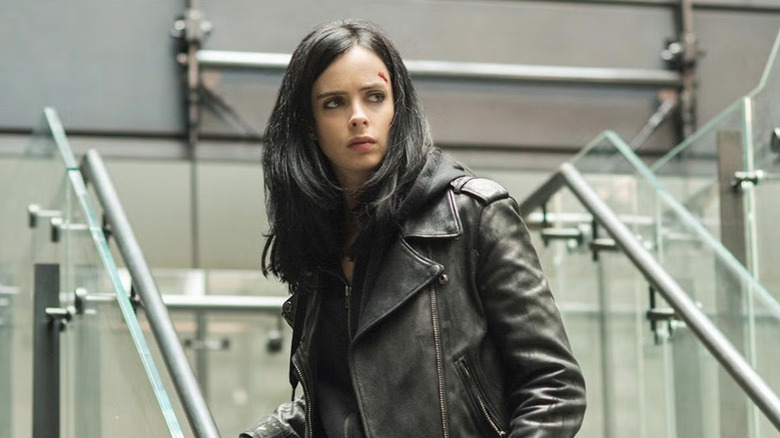 Krysten Ritter with long black hair wearing a black leather motorbike jacket with a black hoodie underneath. There's a small cut on her forehead. She's standing on a glass staircase.