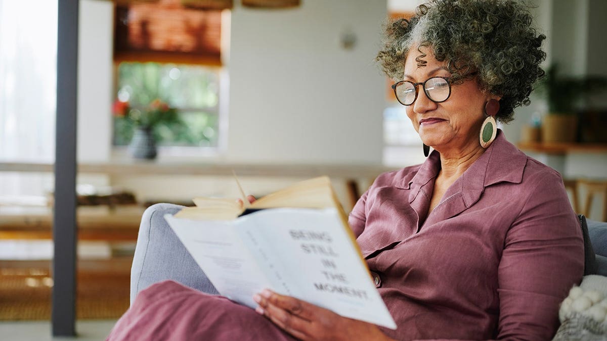 woman reads a book while sitting on a sofa in a living room.