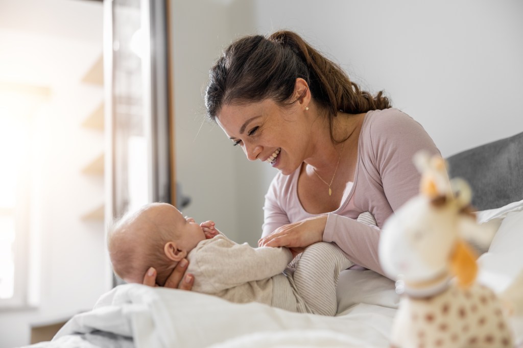 A smiling mother plays lovingly with her baby on a bed in a bright bedroom.