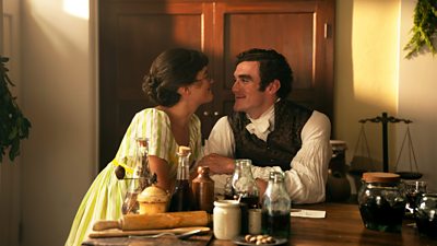 Mary (Ella Bruccoleri) and Tom (Dónal Finn) smile at each other happily as they sit at a table in a warm, domestic home. 
