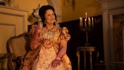Mrs Bennet (Ruth Jones) smiles while holding a glass of champagne. She’s sat in an elegant chair in a warm parlour illuminated by candlelight.