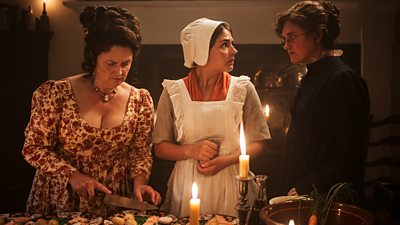 Mrs Bennet (Ruth Jones) and Mary (Ella Bruccoleri) stand either side of a kitchen maid. Mary looks directly at the maid with a stern expression, while Mrs Bennet uses a kitchen knife to prepare food on the table. Warm candlelight illuminates the scene. 