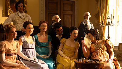 A group of ladies in formal dress sit in a line in a large, lavish living room. Mrs Bennet sits at the end of the group and whispers her confidences to another woman behind a small hand fan. Footmen and a gentleman stand in the background. 
