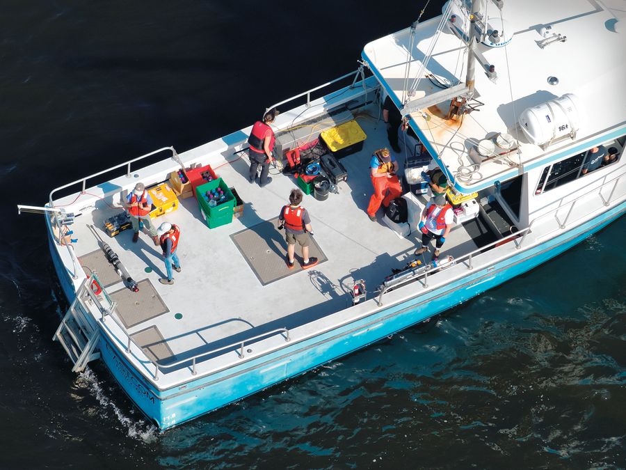 Researchers in safety gear work with instruments and sampling equipment aboard a blue-and-white boat on dark water.