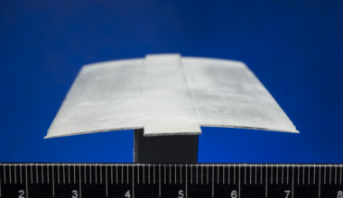A close-up view of a cross section of a rotor blade, also called an airfoil. The thin, gray material is slightly arched and sits atop a black stand against a ruler.