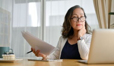 Mature woman in glasses reviewing documents while working at a laptop in a bright home office