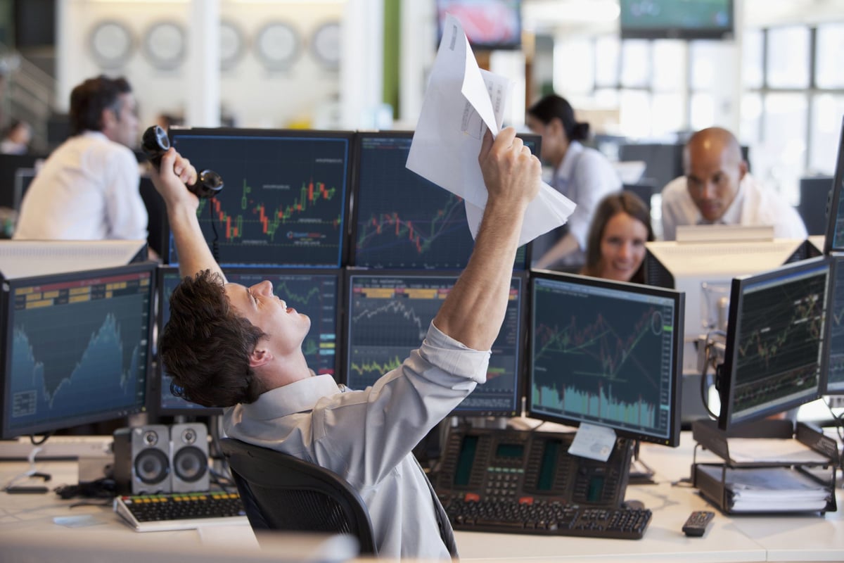 Stock trader celebrates with papers amid monitors displaying financial market charts.