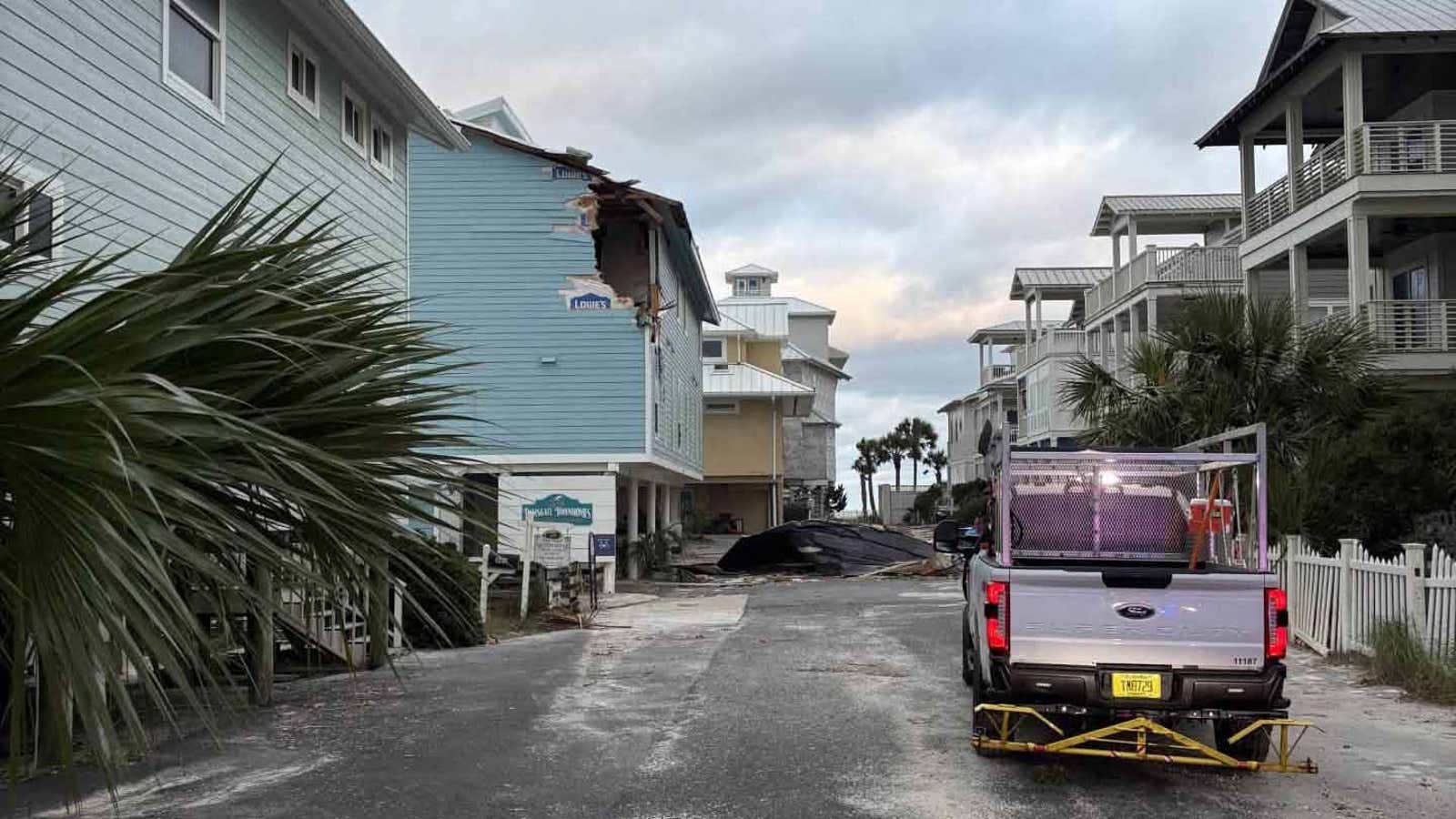 Storm-damaged beach house with torn roof and debris on street, pickup truck parked nearby among coastal townhomes.