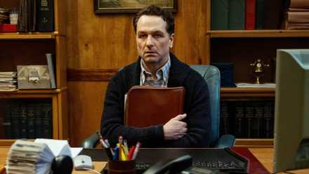 Matthew Rhys as Tom sitting at his office desk while hugging a folder to his chest in Widow's Bay
