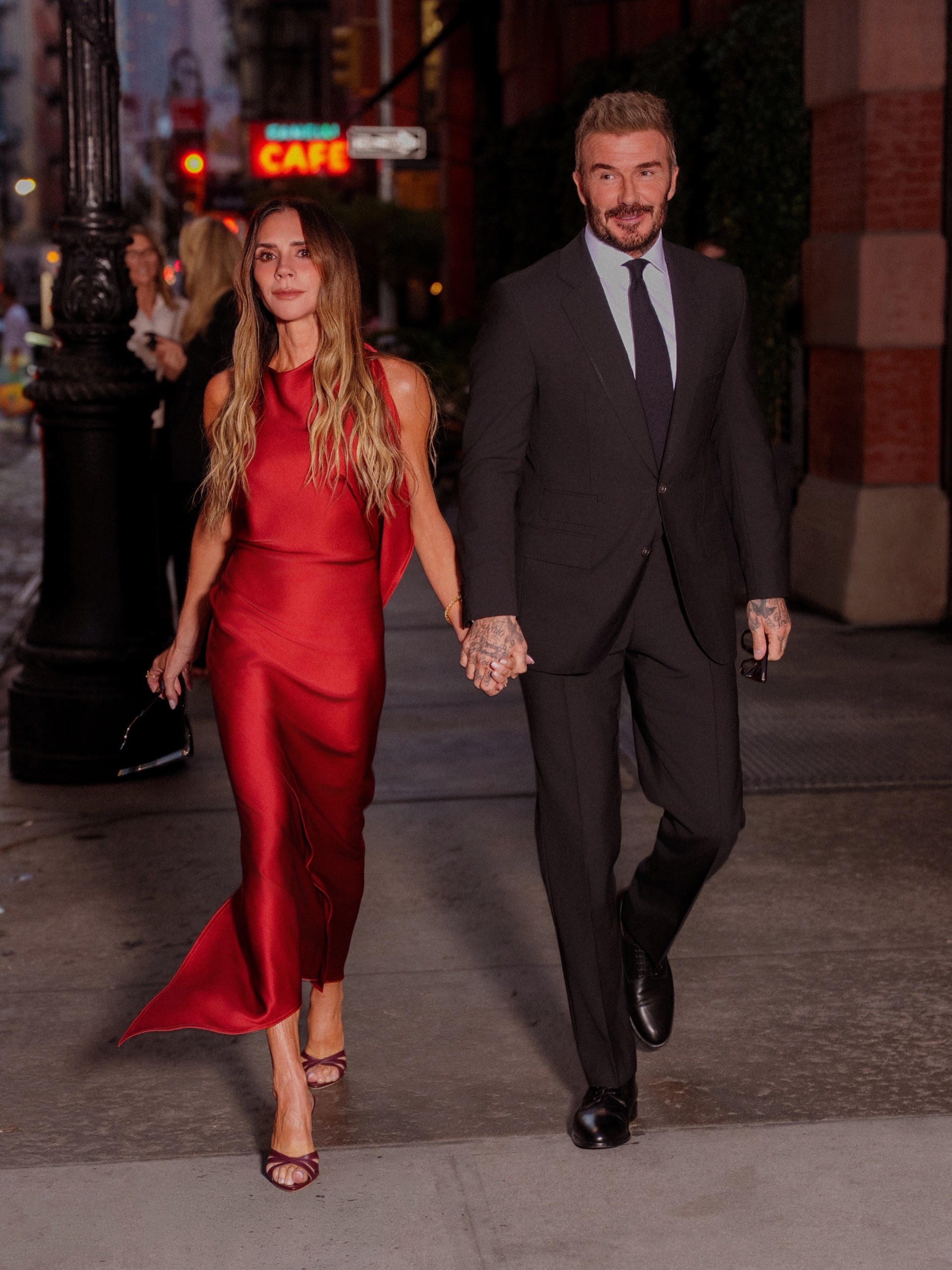Victoria and David Beckham walk down the street holding hands. Victoria wears a red satin dress with plum heels, and David wears a suit.