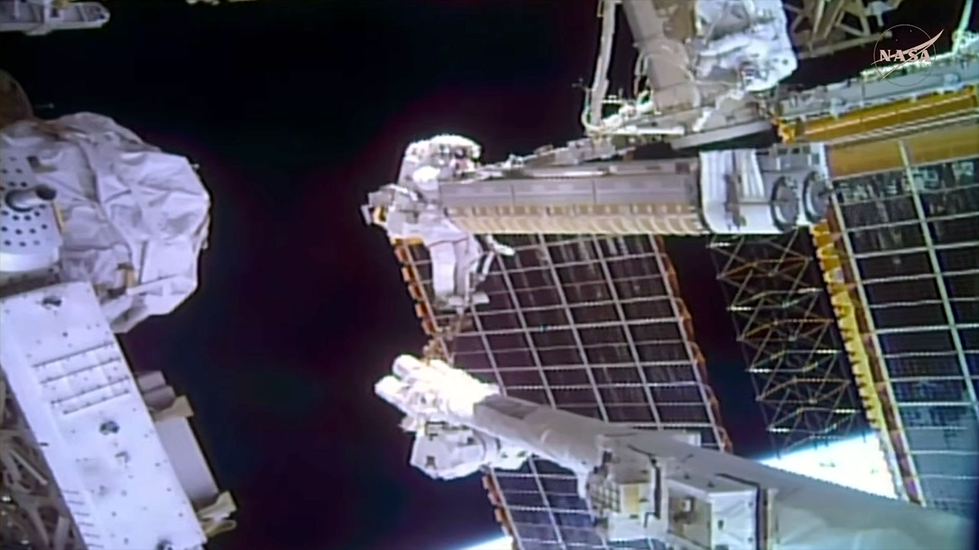 a spacesuited astronaut is seen holding a large piece of cargo while riding at the end of a robotic arm outside of a space station