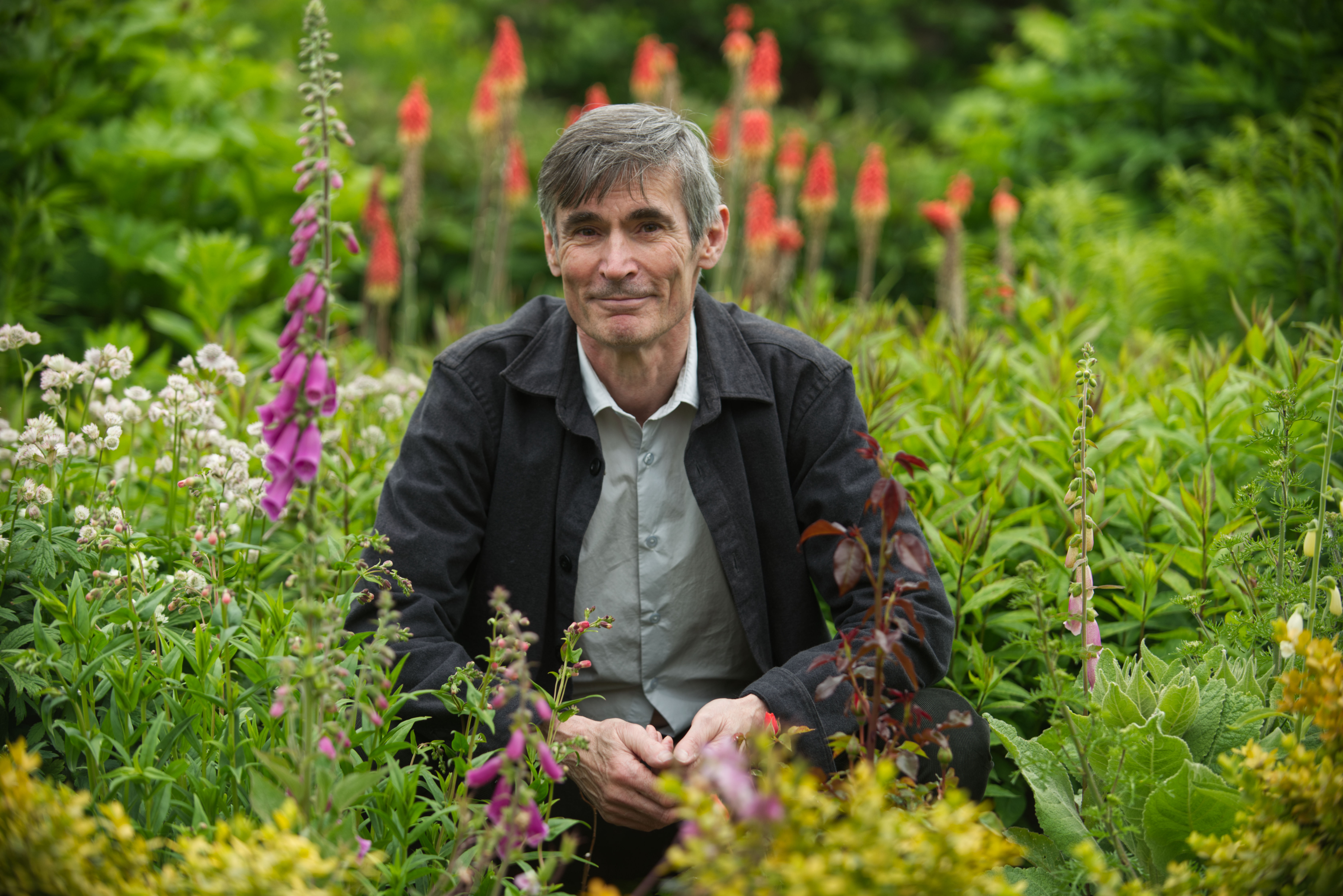 Booker Prize nominee Stephen Brunt kneeling in a lush garden filled with green plants and purple, white, and red flowers.